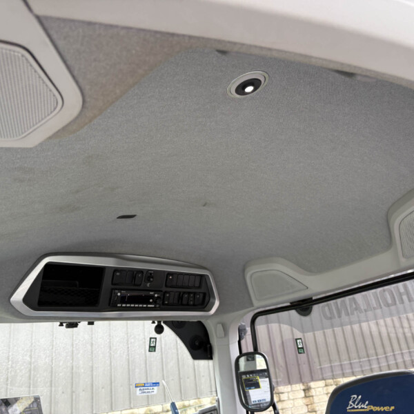 Interior of a truck cab ceiling with overhead console and built‑in speakers seen from the driver's seat edge outside window view of industrial building outside the cab.