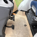 Interior of a vehicle cab with cardboard floor protection, visible pedals and a blue passenger seat beside a gray console.