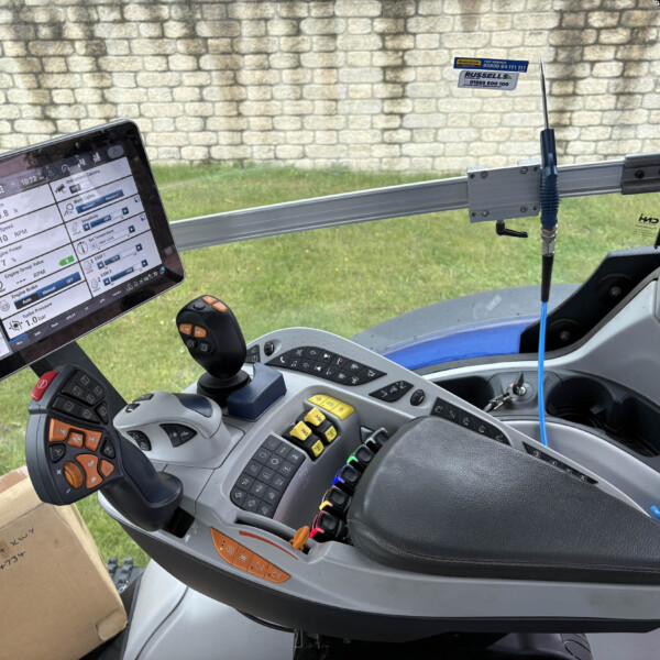 Interior view of a tractor cabin with a mounted touchscreen display showing telemetry, plus a joystick and colored control buttons nearby, with a stone wall outside the window work area.