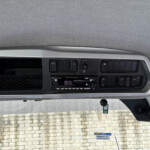 Dashboard panel with multiple switches and a central display, seen from the driver's seat, brick wall visible through the windshield outside.
