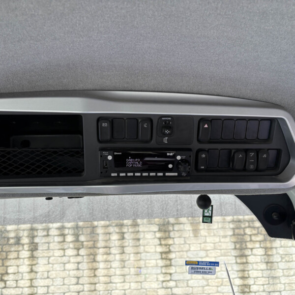 Dashboard panel with multiple switches and a central display, seen from the driver's seat, brick wall visible through the windshield outside.