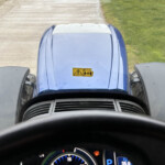 Driver’s view from inside a blue vehicle; steering wheel at the bottom, dashboard gauges visible, hood warning label, road ahead.