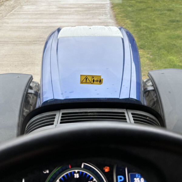 Driver’s view from inside a blue vehicle; steering wheel at the bottom, dashboard gauges visible, hood warning label, road ahead.