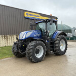 Blue New Holland tractor with oversized tires parked beside a gray industrial building bearing a New Holland Agriculture sign in yellow and blue.
