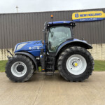 Blue New Holland tractor parked in front of a dark gray corrugated building with a yellow-and-blue New Holland Agriculture sign.