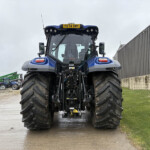 Front view of a blue agricultural tractor with oversized tires, parked in a farm yard near a corrugated metal building.