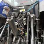 Close-up of a heavy truck’s front hitch and hydraulic linkage with hoses, levers, and warning stickers (wet from rain).