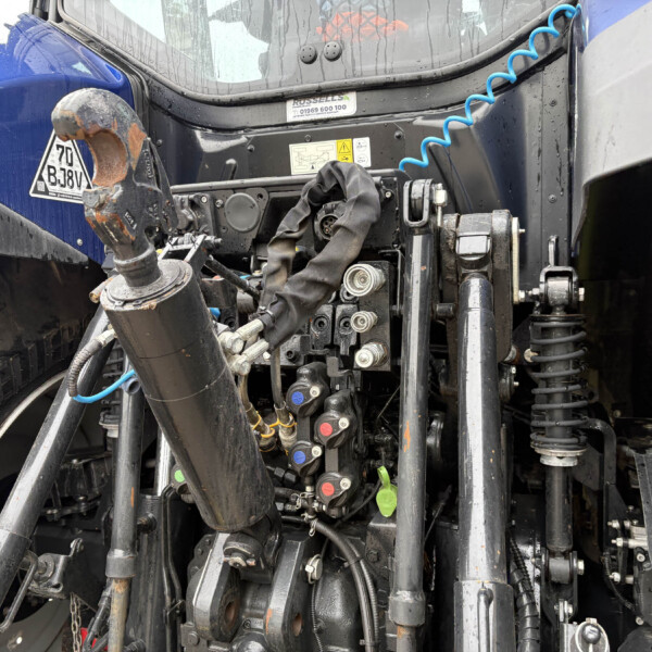 Close-up of a heavy truck’s front hitch and hydraulic linkage with hoses, levers, and warning stickers (wet from rain).