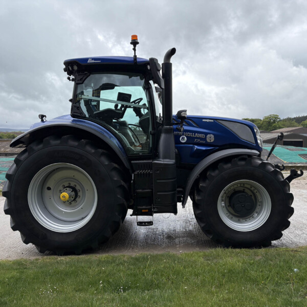 Blue New Holland T7 tractor with large front tires parked on a concrete lot under a cloudy sky, front cabin visible.
