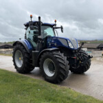 Blue New Holland tractor parked on a farm yard with gray cloudy sky and distant barns in the background; large tires and modern cab visible.