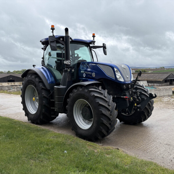 Blue New Holland tractor parked on a farm yard with gray cloudy sky and distant barns in the background; large tires and modern cab visible.
