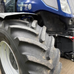 Front section of a blue New Holland tractor with a large muddy front tire, fender, and exposed hydraulic components visible beneath the cab.