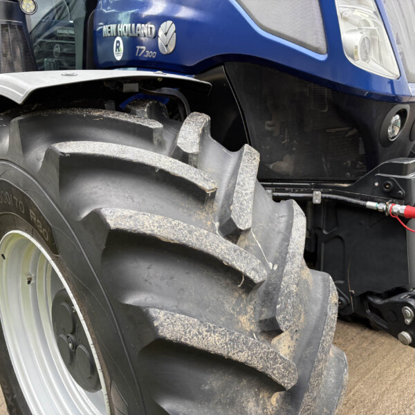 Front section of a blue New Holland tractor with a large muddy front tire, fender, and exposed hydraulic components visible beneath the cab.