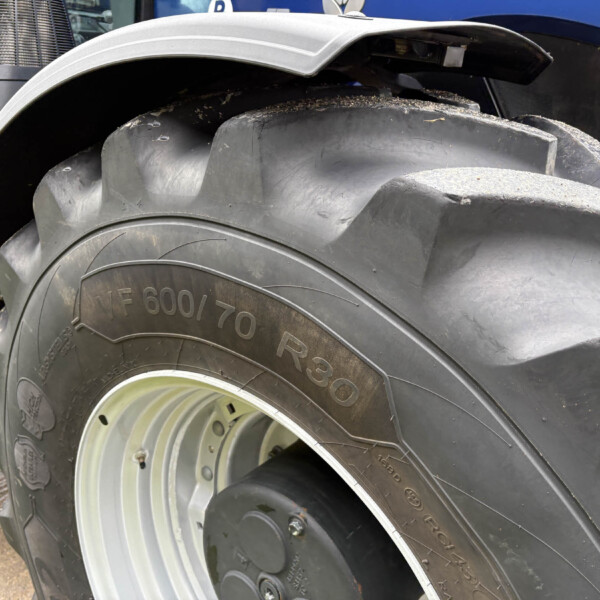 Close-up of a large tractor tire with deep tread and dirt, mounted on a metal rim with a blue vehicle partially visible in the background.