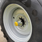 Close-up of a large tractor rear tire and silver rim, showing a yellow hub cap and bolted axle in a rural area.