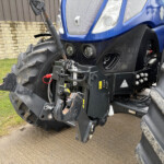 Close-up of the blue tractor's front loader attachment with hydraulic linkages, dirt-covered bucket, and rugged tires near a brick wall.