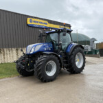 Blue New Holland tractor parked outside a gray industrial building with a yellow-and-blue New Holland sign above it.