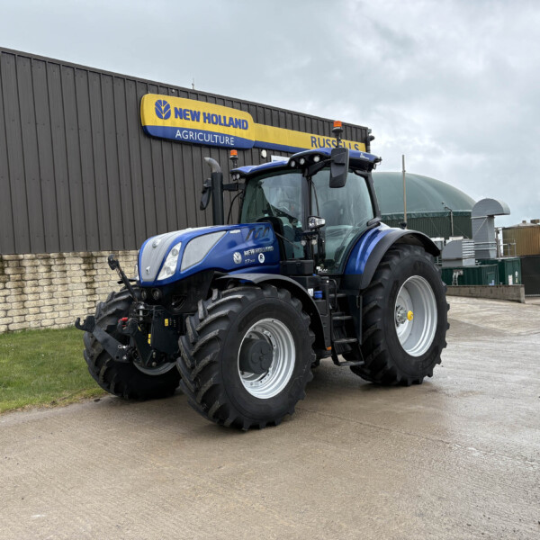 Blue New Holland tractor parked outside a gray industrial building with a yellow-and-blue New Holland sign above it.