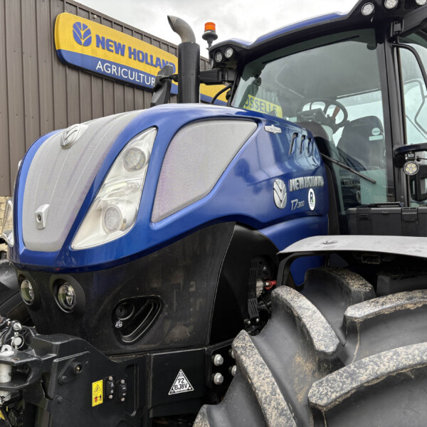 Blue New Holland tractor front view with muddy tires and cab, signage reading New Holland Agriculture in the background.