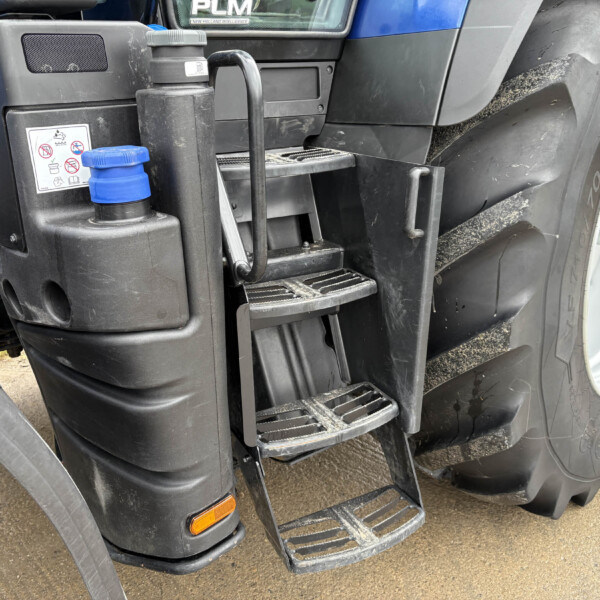 Close-up of a blue-accented tractor front left side showing foot steps, black handrails, a blue cap, and a large dirty tire with a yellow hubcap in the background.