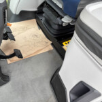 Inside a truck cab, view of the driver’s footwell with rubber pedals, gray carpet, and a cardboard sheet on the floor beside the door.