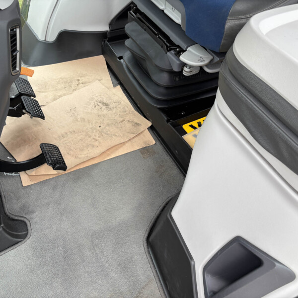 Inside a truck cab, view of the driver’s footwell with rubber pedals, gray carpet, and a cardboard sheet on the floor beside the door.