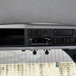 Close-up of a vehicle dashboard showing the radio, buttons, and vents, with a textured headliner above and a stone wall visible through the windshield.