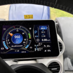 Digital car instrument panel showing speedometer, RPM, and fuel indicators with a steering wheel in view behind it.