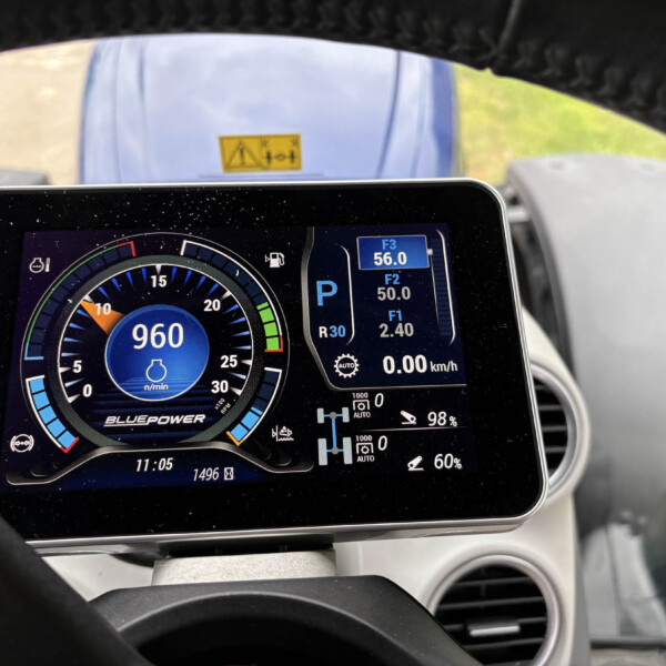 Digital car instrument panel showing speedometer, RPM, and fuel indicators with a steering wheel in view behind it.