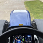 Driver's perspective from inside a blue truck cab, showing the steering wheel and a digital instrument cluster with high-speed readout while road and green grass are visible ahead.