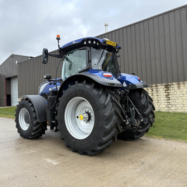 Blue New Holland tractor parked outside a gray industrial building with large tires and a brick foundation.