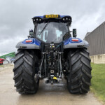 Front view of a blue agricultural tractor with oversized rear tires parked in a yard beside a corrugated metal building under a cloudy sky.