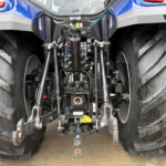 Back of a blue tractor showing hydraulic valves, levers, hoses, and a three-point hitch between muddy, oversized rear tires