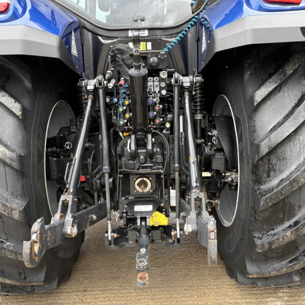 Back of a blue tractor showing hydraulic valves, levers, hoses, and a three-point hitch between muddy, oversized rear tires