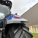 Close-up of a blue tractor front end with windshield, lights, and hydraulic components against a wooden fence backdrop.
