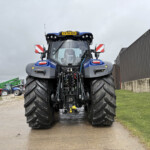 Front view of a large blue agricultural tractor with oversized tires parked on a muddy path beside a corrugated metal building.