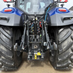 Rear view of a large blue tractor showing exposed hydraulic plumbing, hoses, and oversized tires.