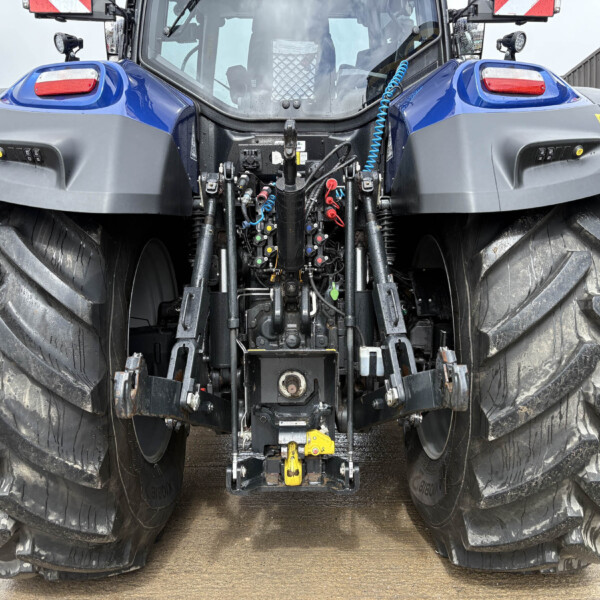 Rear view of a large blue tractor showing exposed hydraulic plumbing, hoses, and oversized tires.