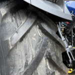 Close-up of a large black tire coated in mud, with parts of a vehicle's frame and blue bodywork visible in the background.