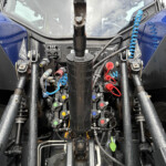 Close-up of a tractor's hydraulic control area with a central black joystick, multiple levers, colored caps, and blue coiled hose, all wet from rain.