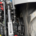 Close-up of exposed hydraulic system and wiring on heavy machinery, showing red caps, blue components, and damp surfaces near a large tire edge.