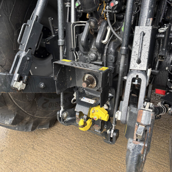 Heavy industrial machinery frame with exposed hydraulic components, hoses, and a yellow safety hook beneath the vehicle's wheel area.