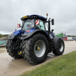 Close-up view of a blue tractor cab with large front tires, standing on a paved surface in an industrial yard, orange beacon on top and red-white roadside mirrors visible on the cabin.