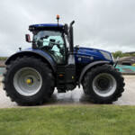 Blue New Holland tractor seen from the side, close to the camera with large front tire and cab, set on a farm yard under a cloudy sky.