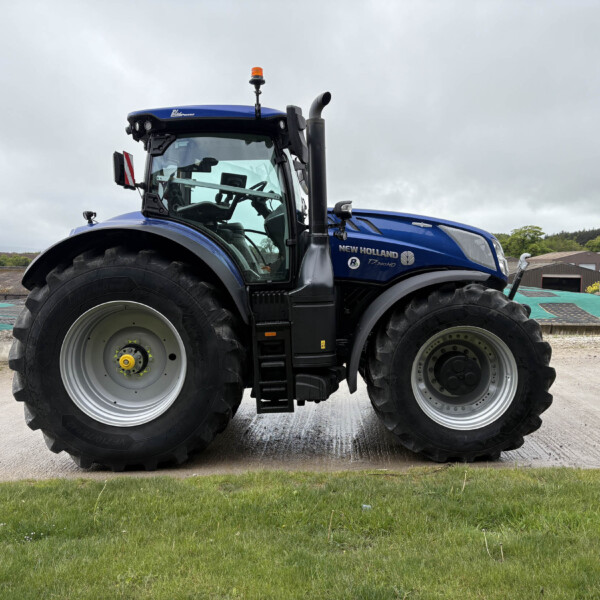 Blue New Holland tractor seen from the side, close to the camera with large front tire and cab, set on a farm yard under a cloudy sky.
