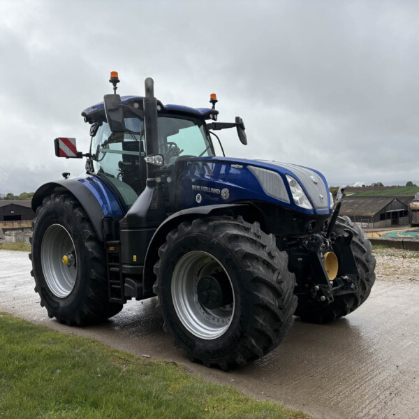 Blue New Holland tractor with oversized tires parked on a farmyard under a cloudy sky, side view showing cabin and orange warning lights.
