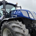 Close-up of a blue New Holland tractor showing the front grille, large tire, and cab window from a low angle.