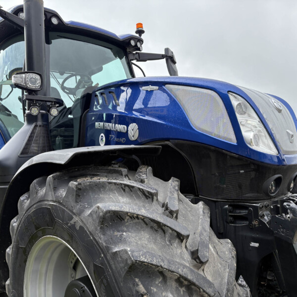 Close-up of a blue New Holland tractor showing the front grille, large tire, and cab window from a low angle.