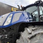 Close-up of a blue New Holland tractor front, showing the grille, headlights, and muddy tires with a metal wall in the background.