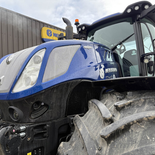 Close-up of a blue New Holland tractor front, showing the grille, headlights, and muddy tires with a metal wall in the background.